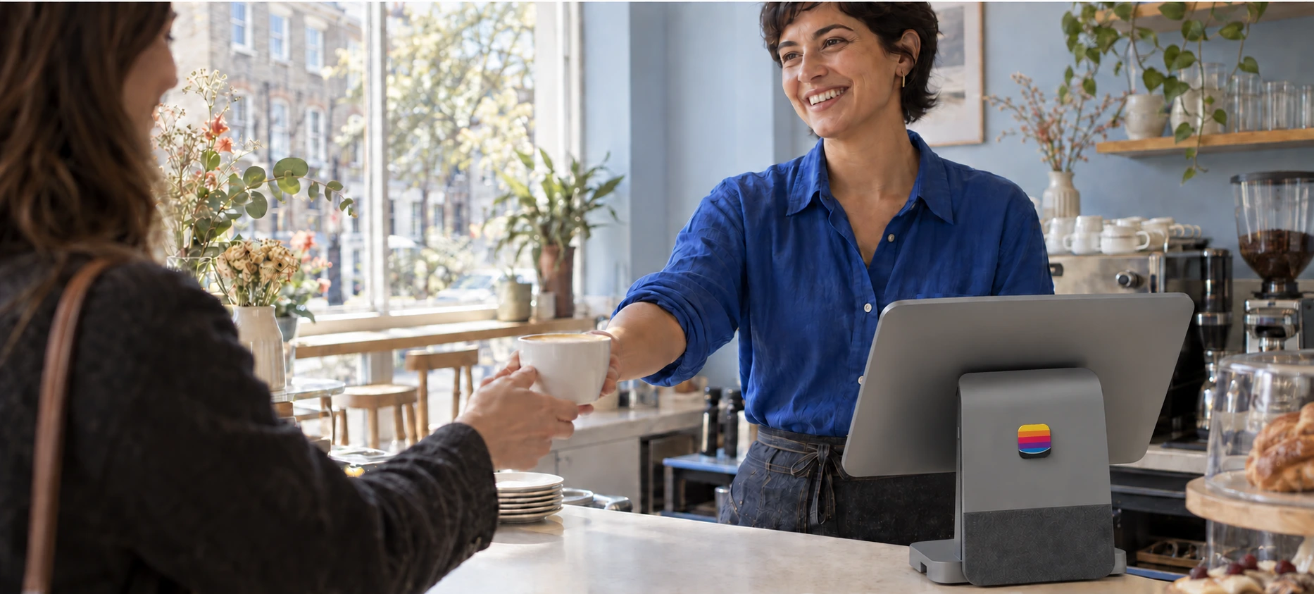 A café owner handing a coffee to a customer beside a Tillable register with a raised rainbow logo mark.